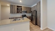 This single-floor residence offers a thoughtfully designed layout that balances comfort and functionality. The kitchen is a standout feature, showcasing rich wood-toned cabinetry with open shelving, granite-look countertops, a peninsula with a built-in sink, and a full suite of stainless steel appliances including a refrigerator, microwave, and electric range. Light wood-style flooring runs throughout the main living areas, lending a clean and cohesive aesthetic.

The open living area is bright and inviting, benefiting from a large window fitted with blinds that allows generous natural light to fill the space. A textured ceiling adds subtle character throughout the common areas. A hallway connects the main living spaces to the private quarters, where bedrooms feature plush wall-to-wall carpeting and windows with outdoor views. At least one bedroom includes a walk-in closet with built-in hanging rods, providing practical storage.

The bathrooms are well-appointed, with one featuring a dual-sink vanity with wood-tone cabinetry, granite countertops, and light wood flooring. A second bathroom offers a tiled tub-and-shower combo, single-sink vanity with matching cabinetry, and coordinating finishes. Overall, the space presents a modern, neutral palette designed for easy living.