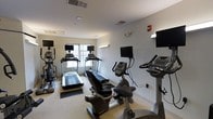This fitness center offers a practical and well-organized space for cardiovascular exercise and general workouts. The room features light-colored walls and a neutral carpeted floor, contributing to a clean and open atmosphere. Natural light filters in through windows along one wall, brightening the space and creating a welcoming environment for exercise.

The facility is equipped with multiple treadmills positioned near the windows, each outfitted with individual monitors for entertainment during workouts. Additional cardio equipment includes stationary upright bikes and a recumbent bike, all from Cardio brand machines, providing a variety of low-impact exercise options. Wall-mounted flat-screen televisions are installed at strategic viewing angles, ensuring visibility from multiple workout stations.

The ceiling features recessed lighting and HVAC vents, maintaining a comfortable climate throughout the space. A fire alarm system is visibly installed, reflecting attention to safety standards. The overall layout is thoughtfully arranged to allow users to move freely between equipment without congestion. This single-floor fitness center is designed to deliver a functional and accessible workout environment, with an emphasis on cardio training supported by modern equipment and adequate entertainment amenities.