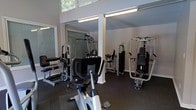 This fitness center is a dedicated, single-floor exercise facility designed to accommodate a comprehensive range of workout needs. The space features high ceilings with an angled roofline that contributes to an open, airy feel, while large windows along the upper walls invite natural light and offer views of surrounding greenery. Structural columns are integrated throughout the room, adding architectural character without compromising the usable floor area. The flooring consists of dark rubber matting, providing both durability and safety for equipment use. The facility is equipped with a solid selection of strength training machines, including multi-use cable systems and dedicated resistance machines targeting various muscle groups, as well as a recumbent stationary bike for cardiovascular training. Overhead fluorescent lighting ensures consistent illumination throughout the space. The neutral gray walls and clean layout create an organized and focused environment. Whether used for strength building, rehabilitation, or general fitness, this well-appointed facility offers users a practical and comfortable setting to pursue their health and wellness goals.