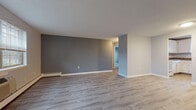 Located at 126 New Britain Ave in Plainville, Connecticut, this single-floor unit offers a clean, updated interior with a cohesive neutral color palette throughout. The living areas are finished with light gray laminate hardwood flooring that extends seamlessly across the space, contributing to a bright and open feel. Walls are painted in soft gray tones, complemented by crisp white baseboard trim, creating a modern yet understated ambiance. Natural light filters in through multiple windows fitted with blinds, enhancing the spaciousness of each room. Baseboard heating units are present throughout, providing efficient climate control, and a wall-mounted air conditioning unit is also available. The kitchen is well-appointed with white raised-panel cabinetry, dark laminate countertops, a double-basin sink, and tile flooring. White appliances include an electric range, dishwasher, and refrigerator, along with ample upper and lower cabinet storage. A dedicated hallway connects the main living area to the bedroom and bathroom. The bedroom features the same gray laminate flooring, a window with a view of surrounding greenery, and generous wall space. The bathroom is neatly finished with wood-look flooring, a white vanity, toilet, and a tub-shower combination, maintaining the clean, functional aesthetic found throughout the unit.