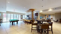 This well-appointed single-floor amenity space offers a diverse range of social and recreational areas, unified by a sophisticated design aesthetic that blends warm wood tones, neutral upholstery, and bold decorative accents. The expansive game and entertainment area anchors the space, featuring a regulation pool table, shuffleboard, and a curved bar with pendant lighting, dark wood cabinetry, built-in appliances, and bar seating. Large windows and glass doors allow generous natural light and outdoor views throughout this central zone, with tile flooring adding a clean, durable finish.

Adjacent lounge areas offer more intimate settings, including a gallery room with Barcelona-style leather chairs, a zebra-print rug, wall-mounted televisions, and a striking grid of framed equestrian artwork. Oversized vintage-style mugshot prints add an eclectic, bold touch to another nook equipped with a shuffleboard table and built-in storage.

The business center features rich blue accent walls, warm wood flooring, built-in workstations with computer terminals, and glass-front cabinetry. A separate lounge near the entrance provides a bright, welcoming sitting area with cream-toned armchairs, patterned wallcoverings, and floor-to-ceiling windows. Throughout, the curated art, layered textures, and varied seating arrangements create a dynamic and inviting communal environment.