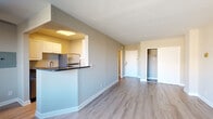 This well-appointed single-floor unit offers a clean, modern aesthetic with warm neutral tones and light-toned hardwood flooring throughout. The open living area is generously sized, benefiting from a large window fitted with horizontal blinds that allows ample natural light to fill the space. A wall-mounted light fixture adds a subtle design touch, while a baseboard radiator provides efficient heating.

The galley-style kitchen is efficiently laid out and features white cabinetry, dark granite countertops, and stainless steel appliances including a refrigerator, gas range, and a range hood. A stainless steel sink with a modern faucet completes the functional workspace. The kitchen connects to the main living and dining area via a pass-through opening, supporting an open and social feel.

The bedroom is a well-proportioned room with generous natural light streaming in through large windows that frame views of surrounding buildings. The room includes a baseboard heating unit and ample wall space. A closet with bifold doors provides practical built-in storage.

The bathroom features classic white tile work, a tub-shower combination with a curtain rod, and a vanity with a mirror. The layout is clean and functional. Throughout the unit, crisp white baseboards and neutral wall tones create a cohesive and inviting atmosphere.