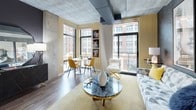 This single-floor urban loft showcases an industrial-chic aesthetic defined by exposed concrete ceilings with visible ductwork, hardwood-style flooring, and an open-concept layout that maximizes both space and natural light. Large, floor-to-ceiling windows wrap around the living and dining area, framing striking city views of surrounding brick buildings and filling the interior with abundant daylight.

The main living area features a cohesive design anchored by warm yellow accent walls, a statement marble-top coffee table with a gold base, and eclectic furnishings. A dining nook with a round table and mustard-toned chairs sits adjacent to the windows, while the living space flows seamlessly toward the sleeping area.

The bedroom is elegantly separated from the living space by sleek sliding glass-panel partitions, providing privacy without sacrificing the open feel. The bedroom walls are dressed in a sophisticated herringbone-patterned textured wallcovering that adds depth and contrast.

The kitchen is well-appointed with white cabinetry, quartz countertops, a classic subway tile backsplash, stainless steel appliances, and matte black hardware throughout. The bathroom continues the clean aesthetic with a white vanity, light tile flooring, and cool grey walls accented by matte black fixtures.