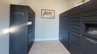 This single-floor parcel delivery room is purpose-built to streamline package management for residents. The space is accessed via a glass-panel exterior door clearly marked for parcel deliveries, allowing couriers direct and efficient entry from the outside. Natural light flows in through a large fixed window alongside the entry door, offering views of the surrounding landscaped exterior and parking area.

At the heart of the room, two tall columns of dark charcoal-toned package lockers face each other, forming a central aisle and providing an extensive array of secure, individually keyed compartments in varying sizes. The lockers are finished in a sleek matte black, lending a modern and orderly aesthetic to the space. Light wood-look luxury vinyl flooring runs throughout, complementing the neutral, light gray wall tones. A framed piece of artwork is mounted on the far wall between the locker banks, adding a refined touch to an otherwise functional environment.

Overhead lighting ensures the space remains well-lit at all times. A security camera is visible near the ceiling, emphasizing safety and accountability. An interior door connects the parcel room directly to the residential corridor, where light-filled hallways lead toward units featuring open kitchens with dark cabinetry, stainless steel appliances, and island seating — suggesting this delivery room is part of a well-appointed residential community.