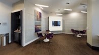 This single-floor commercial space is designed with a polished, contemporary aesthetic that balances functionality with refined style. The entry area centers around a curved reception desk finished in a light stone surface with a dark base, accompanied by upholstered bar-height seating. Textured wave-patterned wall panels add depth and visual interest throughout, complementing the neutral color palette of soft grays and whites.

Pendant linear lighting fixtures are suspended from smooth white ceilings, providing consistent, professional illumination across all areas. The flooring throughout is a rich, dark-toned carpet with a subtle woven pattern, grounding the space with warmth.

A dedicated lounge and meeting area features a round wood-topped table surrounded by plush purple upholstered chairs, alongside a wall-mounted flat-screen display ideal for presentations. Curated large-format artwork adorns the walls, adding a creative dimension to the professional environment.

Floor-to-ceiling glass partitions and doors allow sightlines between zones while maintaining an open, connected feel. Additional seating nooks with patterned accent chairs and a low white cocktail table provide flexible casual meeting options. A private room with a frosted glass door offers an enclosed space for more confidential use.
