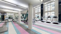 Located at 715 US Highway 46, this contemporary two-floor fitness center offers a well-appointed and expansive workout environment designed for a full range of exercise activities. The main floor is the larger of the two levels, featuring soaring double-height ceilings and floor-to-ceiling windows that flood the space with natural light and offer views of an outdoor pool area. A bold, multi-colored striped carpet in pink, teal, gray, and white defines the cardio zone, which is equipped with a full row of treadmills, ellipticals, stationary bikes, and Echelon smart equipment. Decorative patterned columns add a distinctive architectural element throughout. Full-length mirrors line the walls, enhancing the sense of depth and space. A dedicated functional training zone features artificial turf flooring and houses free weights, squat racks, battle ropes, sled equipment, and air bikes — ideal for high-intensity training. A striking teal spiral staircase connects to the upper level, which provides additional workout space and overlooks the main floor below. Linear LED lighting runs throughout both floors, ensuring consistent, well-distributed illumination. The overall design balances energetic aesthetics with practical, high-performance fitness infrastructure.