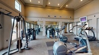 Located at 5701 Saratoga Blvd in Corpus Christi, Texas, this well-appointed fitness center offers a comprehensive workout environment designed to accommodate a wide range of exercise needs. The single-floor facility features soaring ceilings with coffered detailing and recessed lighting, creating a bright and open atmosphere that enhances the sense of space throughout.

The workout floor is equipped with an extensive lineup of professional-grade Precor cardio and strength-training machines, including treadmills, ellipticals, stationary bikes, stair climbers, and multi-station cable machines. Equipment is arranged to allow comfortable circulation and easy access throughout the space. Carpet flooring with rubber mat overlays beneath the equipment provides both durability and underfoot comfort during workouts.

Floor-to-ceiling mirrors line the walls, visually expanding the room and allowing users to monitor their form during exercise. Large windows and glass doors on multiple sides of the facility flood the space with natural light while offering views of the surrounding outdoor landscape, including palm trees characteristic of the South Texas setting. The glass doors also provide direct outdoor access, adding to the facility's open, airy feel.

The overall ambiance is clean, professional, and well-maintained, making this fitness center a functional and inviting space suitable for a full range of fitness routines.