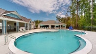 Located at 4605 Sidbury Crossing Dr in Castle Hayne, NC, this two-floor community amenity center offers a well-appointed outdoor retreat designed for relaxation and recreation. The centerpiece is a generously sized freeform swimming pool with a shallow wading area and a separate kiddie/splash pool nearby, providing options for swimmers of all ages. The pool features gentle entry steps and handrails, with depth markers clearly visible along the edges.

Surrounding the pool is an expansive concrete sundeck furnished with lounge chairs and patio seating shaded by vibrant teal umbrellas, creating a welcoming atmosphere for outdoor leisure. The deck connects seamlessly to the main clubhouse, which features large sliding glass doors that open the interior to the outdoors, enhancing the indoor-outdoor flow. The clubhouse is designed with a covered porch and ceiling fans to provide comfort during warmer months.

Architecturally, the facility includes a secondary structure with a distinctive metal roof in a complementary coastal style, alongside a gated pool house with restroom and utility access. The entire complex is enclosed by a black iron fence and is framed by tall, mature pine and hardwood trees that provide natural privacy and a peaceful, wooded ambiance. This amenity center serves as a vibrant hub for community activity in a natural North Carolina setting.