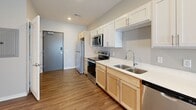This single-floor unit offers a thoughtfully designed open-concept layout that seamlessly connects the kitchen and living area, creating a bright and functional living space. The kitchen is well-appointed with stainless steel appliances, including an electric range, over-the-range microwave, dishwasher, and refrigerator. A dual-basin stainless steel sink sits beneath upper white shaker-style cabinetry, complemented by warm-toned lower cabinets and clean white countertops. Recessed lighting illuminates the kitchen, enhancing its polished, modern feel. Wood-look plank flooring extends throughout the main living areas, adding warmth and visual continuity. The open living area is generously proportioned, benefiting from natural light through vertical blinds and additional windows. Neutral gray walls and white trim provide a clean, versatile backdrop throughout. A utility panel is conveniently accessible in the hallway area, which also connects to additional rooms. The bathroom features a full walk-in shower with a curved curtain rod, a white surround enclosure, and polished chrome fixtures, complemented by matching wood-look flooring. The layout, as seen in the 3D overview, is efficient and well-organized, making excellent use of the available space.
