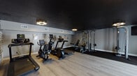 This dedicated fitness center is designed to support a comprehensive workout experience. The space is defined by a bold aesthetic, featuring a dark matte black ceiling that contrasts sharply with the light gray walls and smooth light wood laminate flooring. Unique gear-shaped pendant light fixtures provide warm, focused illumination throughout the room.

The cardio zone is generously stocked with multiple treadmills, elliptical trainers, and stationary bikes—both upright and recumbent—offering a variety of options for cardiovascular exercise. A dedicated strength training area features a multi-function cable machine and a weight bench, positioned on protective rubber flooring matting.

A separate free weights area includes a fully stocked dumbbell rack and a weight bench, situated near the entrance, which consists of double glass-panel doors that allow natural light to fill that corner of the room. A cork bulletin board is mounted nearby for informational postings.

Adding a distinctive visual element, one wall is clad in a rustic wood-plank accent treatment, giving the space texture and character. A rack of colorful medicine balls and resistance bands rounds out the functional equipment offerings. Framed artwork is displayed throughout, contributing to a polished and motivating atmosphere.