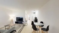 This well-organized single-floor residence offers a thoughtful layout that maximizes every square foot across its four distinct areas: a living room, bedroom, bathroom, and entrance. The open living and dining area is bright and minimalist, featuring light-toned ceramic tile flooring that runs throughout the home. The living space accommodates a seating area with a sofa and flat-screen TV, while a glass-top dining table with modern black chairs defines the dining zone nearby. A compact kitchen station with a microwave and appliances is neatly integrated into the space. The spacious primary bedroom comfortably fits multiple beds and is brightened by a wide window with vertical blinds. A built-in mirrored sliding-door closet enhances the sense of space while providing ample storage. The bathroom features a full tub-shower combination with clean beige tile surround, a chrome showerhead, and matching tile flooring. Throughout the home, white walls and recessed and overhead lighting contribute to a bright, airy ambiance. The layout flows logically from the entrance through the common areas and into the private bedroom, making efficient use of the available footprint.