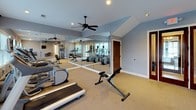 This fitness center is a thoughtfully designed workout facility spread across a single floor, offering a comprehensive range of equipment to support diverse exercise routines. The space is finished with neutral gray walls and carpeted flooring, creating a clean and functional atmosphere throughout.

The cardio area is well-stocked with multiple treadmills and elliptical machines, each equipped with individual display screens for tracking workout metrics. A dumbbell rack provides a full selection of free weights, complemented by kettlebells and a stability ball for varied training options. A cable cross machine rounds out the strength training offerings, supporting a full range of resistance exercises.

Natural light flows in through several windows fitted with horizontal blinds, keeping the space bright and inviting. A large floor-to-ceiling mirror along one wall visually expands the room and allows users to monitor their form during exercise. A ceiling fan and recessed lighting ensure comfort and visibility at any time of day.

The facility includes a wall-mounted television, a direct exterior entrance through glass-panel doors, and posted fitness rules and operating hours for user guidance. The vaulted ceiling in a portion of the space adds an open, airy quality. Overall, this fitness center provides a well-rounded and comfortable environment for achieving a wide variety of health and wellness goals.