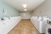 Located at 1410 Hartman Pl in Manhattan, Kansas, this single-story shared laundry facility offers a well-organized and efficiently designed space for residents. The room is generously proportioned, with rows of white top-loading washers and front-loading dryers lining both sides of a wide central aisle, providing ample capacity for multiple users at once. The machines are equipped with digital payment interfaces for convenient operation, and instructional "Easy Washing" guides are posted throughout the room for user reference. Laundry guidelines from the property management are also prominently displayed near the entrance.

The interior features warm-toned ceramic tile flooring that runs the length of the room, contributing to easy maintenance and a clean aesthetic. The walls are painted in soft light blue and gray tones, creating a calm and neutral atmosphere. Flush-mounted fluorescent light fixtures provide consistent overhead illumination, while a window at the far end of the room allows natural light to supplement the artificial lighting. An HVAC vent is positioned near the entrance to maintain comfortable indoor temperatures year-round. The space is uncluttered and functional, with a small utility station centrally located along one wall. Access to an adjacent area is available through a door near the entrance.