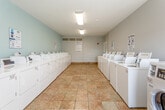 Located at 1410 Hartman Pl in Manhattan, Kansas, this single-story shared laundry facility offers a well-organized and efficiently designed space for residents. The room is generously proportioned, with rows of white top-loading washers and front-loading dryers lining both sides of a wide central aisle, providing ample capacity for multiple users at once. The machines are equipped with digital payment interfaces for convenient operation, and instructional "Easy Washing" guides are posted throughout the room for user reference. Laundry guidelines from the property management are also prominently displayed near the entrance.

The interior features warm-toned ceramic tile flooring that runs the length of the room, contributing to easy maintenance and a clean aesthetic. The walls are painted in soft light blue and gray tones, creating a calm and neutral atmosphere. Flush-mounted fluorescent light fixtures provide consistent overhead illumination, while a window at the far end of the room allows natural light to supplement the artificial lighting. An HVAC vent is positioned near the entrance to maintain comfortable indoor temperatures year-round. The space is uncluttered and functional, with a small utility station centrally located along one wall. Access to an adjacent area is available through a door near the entrance.