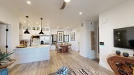This thoughtfully designed single-floor residence combines modern aesthetics with functional living spaces. The heart of the home is an open-concept kitchen and dining area, anchored by a striking white shaker-style kitchen with glass-front upper cabinets, a large center island topped with light stone countertops, and stainless steel appliances including a French door refrigerator. Industrial-style pendant lights hang above the island, while a wall-mounted range hood adds a bold focal point. Light oak hardwood flooring flows seamlessly through the kitchen and into the dining area, where a live-edge wood dining table serves as a natural centerpiece. The living area features sliding glass doors that open to a balcony with views of palm trees, filling the space with natural light. Bedrooms are finished with plush carpeting, ceiling fans, and curated artwork, offering a calm and restful atmosphere. The primary bathroom impresses with a dual-sink vanity featuring a backlit mirror, sleek matte black fixtures, white marble-look countertops, and a glass-enclosed walk-in shower with a rainfall showerhead. Additional bathrooms echo the same refined palette with floating white vanities and large-format tile flooring. Throughout the residence, crisp white walls, recessed lighting, and cohesive design elements create a clean, contemporary ambiance.