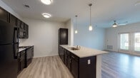 This well-designed single-floor residence offers a thoughtfully laid-out open-concept floor plan that seamlessly connects the kitchen and living areas. The kitchen is a standout feature, showcasing dark espresso cabinetry paired with granite-look countertops, a subway tile backsplash, and black appliances including an electric range and over-the-range microwave. A peninsula with a stainless steel sink and elegant pendant lighting above it creates a natural divide between the kitchen and the open living space, which is finished with light wood-look flooring throughout. The living area is bright and airy, benefiting from natural light and access to an outdoor area through glass French doors. A ceiling fan adds both style and function to this space. The bedroom is a comfortable retreat featuring the same wood-look flooring, a ceiling fan, and a window with outdoor views that fill the room with natural light. The bathroom is clean and contemporary, appointed with large-format tile flooring, a tub and shower combination with classic white subway tile surround, and a dark vanity with a granite-look countertop. The overall design palette of neutral walls, dark cabinetry, and warm wood-look flooring creates a cohesive, modern ambiance throughout the entire unit.