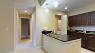 This single-floor residence offers a thoughtfully designed layout that blends functionality with modern finishes. The kitchen serves as a central highlight, featuring striking dark granite countertops that extend across a peninsula-style breakfast bar, providing both prep space and a casual dining option. A double stainless steel sink is neatly integrated into the peninsula, accompanied by stylish cylindrical pendant lights that hang overhead, adding a contemporary touch to the space. Rich, dark-stained cabinetry lines the kitchen walls, offering ample storage while complementing the bold countertop surfaces. Recessed lighting throughout ensures the space remains bright and well-lit. A dedicated in-unit laundry alcove is conveniently tucked adjacent to the kitchen, housing a stacked washer and dryer unit behind louvered doors for a clean, discreet look. The warm, neutral-toned walls carry through from the kitchen into a carpeted hallway, which connects to the remaining rooms of the residence. The light carpeting softens the overall aesthetic and adds a sense of warmth and comfort. The open sightlines between the kitchen and hallway create an airy, connected feel throughout this well-appointed single-floor home.