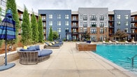 Located at 117 N Oak St in Wichita, Kansas, this contemporary residential community showcases a striking multi-story exterior finished in a sophisticated palette of slate blue siding, warm brick, and light gray panels. The architecture features abundant private balconies and large windows throughout, reinforcing the connection between interior living spaces and the vibrant outdoor environment below.

At the heart of the property lies an expansive courtyard amenity area designed for both relaxation and social engagement. A generously sized swimming pool with a connected spa serves as the central focal point, surrounded by a broad concrete pool deck furnished with blue lounge chairs and circular wicker seating. Adjacent to the pool, a covered pergola structure with wood-slat roofing houses hanging egg-style swing chairs, providing a shaded retreat with an inviting, resort-like atmosphere.

A separate landscaped lawn area offers open green space framed by neatly trimmed shrubs, raised stone planter beds, and accent lighting bollards. An additional courtyard zone features an outdoor dining area with a grill station beneath a pergola canopy strung with bistro lights, ideal for communal gatherings. Manicured landscaping, tiered retaining walls, and thoughtfully placed greenery unify the outdoor spaces into a cohesive and well-appointed community environment.
