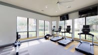 This fitness center offers a comprehensive workout environment thoughtfully designed to support a wide range of exercise routines. The space is divided into distinct functional zones, beginning with a bright, windowed cardio area lined with large floor-to-ceiling windows on multiple sides. These expansive windows flood the room with natural light and provide open views of the surrounding landscaped grounds and outdoor pool area, creating an energizing atmosphere during workouts. The cardio zone is outfitted with multiple treadmills, elliptical machines, and a rowing machine, each equipped with individual monitors. Ceiling fans and recessed lighting contribute to a comfortable environment throughout.

A dedicated strength training area features a full rack of dumbbells, a weight bench, and a multi-functional cable machine system, complemented by wall-to-wall mirrors that enhance the sense of space and allow for form monitoring. An additional functional fitness zone provides stability balls, medicine balls, kettlebells, and resistance accessories for dynamic and flexibility-focused training.

The flooring throughout combines durable carpet tiles in a neutral, patterned design with hardwood-style accents beneath the cardio equipment. Wall-mounted flat-screen televisions are positioned in both the cardio and strength areas. The overall design is clean, modern, and purposefully laid out to accommodate a variety of fitness goals in a well-lit, welcoming setting.