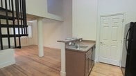 This two-story space showcases a thoughtfully designed interior with warm hardwood floors that run throughout, adding character and a sense of continuity to the layout. The kitchen area features a compact setup with a stainless steel sink, brown cabinetry with under-counter storage, and a laminate countertop that extends into a small breakfast bar overhang — a practical touch for casual dining or additional workspace. A structural column anchors the open floor plan, lending an industrial character to the space. The upper level is partially visible above, with the lofted overhang creating a dynamic sense of vertical dimension and adding architectural interest. Neutral cream-toned walls keep the space feeling bright and open, while a standard exterior door provides convenient access. The overall ambiance is utilitarian yet inviting, with the natural wood flooring serving as the standout design element that ties the space together. The open layout allows for flexible furniture arrangements, making it well-suited for a variety of uses across both floors.