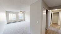 Unit 104 is a single-floor residence designed with a clean, functional layout and a neutral color palette throughout. The living area is generously proportioned, featuring plush wall-to-wall carpeting, a ceiling fan, and a wide window that allows natural light to fill the space. A baseboard heating unit runs along the exterior wall, providing efficient warmth. The room is anchored by a textured popcorn ceiling, consistent with the rest of the unit.

The entryway area transitions into a separate zone with light wood-look luxury vinyl plank flooring, offering a durable and easy-to-maintain surface near the front door. A flush-mount ceiling light and baseboard heater are also present here.

The galley-style kitchen is well-appointed with white cabinetry, gray laminate countertops, a stainless steel sink, a black electric range, and a dishwasher. The light flooring continues into this space, creating a cohesive feel.

The bedroom features soft carpeting, a ceiling fan, a large window for natural light, and a sliding-door closet with a rod and shelf for storage. The bathroom includes a full tub and shower combo with a curved curtain rod, light vinyl flooring, and a vanity with a gray countertop.