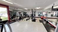This single-level fitness center offers a comprehensive workout environment designed to accommodate a variety of exercise routines. The space is organized into distinct functional zones, with rubber mat flooring strategically placed beneath strength training equipment to contrast the light-toned tile throughout the rest of the room. Floor-to-ceiling mirrors line multiple walls, visually expanding the space and allowing users to monitor their form during workouts.

The cardio section features several treadmills, elliptical trainers, and a stationary bike, including equipment from recognized brands such as Precor. The strength training area is anchored by a multi-station cable machine, a lat pulldown apparatus, and free weight accessories including barbells and medicine balls. A wall-mounted pull-up station adds further versatility for bodyweight training.

Large windows framed in bold red trim run along one wall, flooding the space with natural light and offering views of the surrounding outdoor area. Overhead fluorescent lighting supplements illumination throughout. A textured ceiling and wall-mounted air conditioning units complete the practical, functional design of this well-appointed fitness facility.