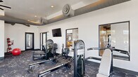 This contemporary fitness center is designed to support a comprehensive workout experience within a single, open-plan floor. The space is defined by crisp white walls, a dark speckled rubberized floor, and an industrial-style ceiling featuring exposed ductwork and cylindrical ventilation units that add a bold architectural character. Recessed lighting and a ceiling fan maintain a comfortable environment throughout.

Large floor-to-ceiling windows line multiple walls, allowing abundant natural light to flood the space and offering views of the surrounding exterior. A full-length mirror wall visually expands the room while providing practical utility for form checking during exercise.

The cardio zone is well-appointed with multiple treadmills and a stair climber, all positioned to take advantage of the natural light from the expansive windows. The strength training area includes a multi-station cable machine, a variety of resistance machines, free weights with a dedicated dumbbell rack, adjustable weight benches, rowing machines, and a medicine ball rack. A stability ball rounds out the functional fitness offerings.

A wall-mounted flat-screen television is installed in one section of the facility, and interior windows provide sightlines into an adjacent lounge or common area, enhancing the sense of connectivity within the broader amenity space.