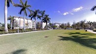 This outdoor common area is part of a multi-story residential community that reflects a classic Florida architectural aesthetic. The grounds are characterized by a wide, open expanse of neatly manicured lawn that provides a generous sense of space and openness. Towering palm trees are strategically positioned throughout the area, lending a tropical character and natural shade to the environment.

The surrounding residential buildings are several stories tall, featuring light-toned exterior finishes consistent with warm-climate construction. Their uniform design and organized layout contribute to a cohesive, well-planned community feel. Walkways and pedestrian paths weave through the landscaped grounds, connecting various parts of the community and encouraging outdoor activity.

Parking areas are visible along the periphery, keeping vehicle traffic separated from the primary green space. Mature shrubs and ornamental plantings along the building edges add layers of greenery and visual interest. The entire setting benefits from abundant natural sunlight under a clear blue sky, creating a bright and welcoming outdoor environment typical of a well-maintained Florida residential community.