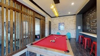 The Retreat's recreational space combines rustic and contemporary design elements to create an engaging, social environment. The main area centers around a full-sized pool table with bold red felt and a reclaimed wood frame, positioned beneath a striking cluster pendant light fixture with amber-toned globes that serve as a dramatic focal point. The flooring features dark hardwood, contrasting beautifully with the textured natural stone brick walls that lend a warm, industrial character to the space. Dark wood crown molding frames the ceiling, adding architectural definition throughout. A slatted wood partition wall with vertical oak slats elegantly divides the billiards area from an adjacent lounge room, which is partially visible and features its own seating and a whiteboard, suggesting use as a meeting or study area. Contemporary teal accent chairs are positioned near the pool table, adding a playful pop of color. Recessed lighting complements the pendant fixture, ensuring the space is well-illuminated. A decorative Louisville Slugger poster adds a sporty, energetic touch to the overall ambiance, making this a well-appointed space designed for leisure and social interaction.
