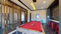 The Retreat's recreational space combines rustic and contemporary design elements to create an engaging, social environment. The main area centers around a full-sized pool table with bold red felt and a reclaimed wood frame, positioned beneath a striking cluster pendant light fixture with amber-toned globes that serve as a dramatic focal point. The flooring features dark hardwood, contrasting beautifully with the textured natural stone brick walls that lend a warm, industrial character to the space. Dark wood crown molding frames the ceiling, adding architectural definition throughout. A slatted wood partition wall with vertical oak slats elegantly divides the billiards area from an adjacent lounge room, which is partially visible and features its own seating and a whiteboard, suggesting use as a meeting or study area. Contemporary teal accent chairs are positioned near the pool table, adding a playful pop of color. Recessed lighting complements the pendant fixture, ensuring the space is well-illuminated. A decorative Louisville Slugger poster adds a sporty, energetic touch to the overall ambiance, making this a well-appointed space designed for leisure and social interaction.