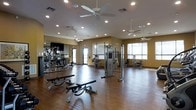 This fitness center offers a generously sized, single-floor workout space designed to accommodate a full range of exercise needs. The room features rich hardwood-style flooring that spans the entire area, providing a clean and durable surface for various training activities. Warm yellow-toned walls create an energizing atmosphere, while multiple large windows along the perimeter flood the space with natural light, enhancing the open and airy feel.

The ceiling is fitted with several ceiling fans and recessed lighting, ensuring comfortable air circulation and consistent illumination throughout. A full wall of mirrors along one side visually expands the room and allows users to monitor their form during workouts.

The facility is equipped with a variety of strength-training machines, including cable and pulley systems, as well as free-weight benches and additional resistance equipment. The layout is thoughtfully organized, allowing for smooth movement between stations without overcrowding. The combination of functional equipment, bright natural light, and well-maintained finishes makes this fitness center a practical and welcoming space for exercise.