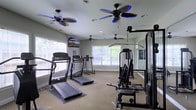 This fitness room offers a comprehensive workout environment designed to support a range of exercise routines. The space is outfitted with multiple treadmills positioned near large windows that allow generous natural light to fill the room, creating an inviting and energizing atmosphere. The windows also provide views of surrounding greenery, enhancing the overall workout experience.

For strength training, a multi-station cable and weight machine is positioned along one side of the room, complemented by a mirrored wall that allows users to monitor their form. The mirrors also contribute to a sense of openness within the space. Carpeted flooring runs throughout, providing a comfortable surface underfoot.

Overhead, multiple ceiling fans — including two with distinctive blue-accented blades — ensure consistent airflow and comfort during workouts. A wall-mounted television is also present, offering entertainment options for cardio sessions. Recessed lighting supplements the natural light, keeping the space bright and well-illuminated at all hours. Overall, the fitness room is practical, well-ventilated, and thoughtfully arranged to accommodate a variety of exercise needs.