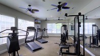 This fitness room offers a comprehensive workout environment designed to support a range of exercise routines. The space is outfitted with multiple treadmills positioned near large windows that allow generous natural light to fill the room, creating an inviting and energizing atmosphere. The windows also provide views of surrounding greenery, enhancing the overall workout experience.

For strength training, a multi-station cable and weight machine is positioned along one side of the room, complemented by a mirrored wall that allows users to monitor their form. The mirrors also contribute to a sense of openness within the space. Carpeted flooring runs throughout, providing a comfortable surface underfoot.

Overhead, multiple ceiling fans — including two with distinctive blue-accented blades — ensure consistent airflow and comfort during workouts. A wall-mounted television is also present, offering entertainment options for cardio sessions. Recessed lighting supplements the natural light, keeping the space bright and well-illuminated at all hours. Overall, the fitness room is practical, well-ventilated, and thoughtfully arranged to accommodate a variety of exercise needs.