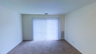 This single-floor space presents a straightforward, functional layout with a clean and neutral aesthetic throughout. The main living area is finished with wall-to-wall carpet in a warm, understated tone, providing a comfortable underfoot feel and a versatile base for various furnishings and decor styles. The walls are painted in a light, neutral hue that contributes to a bright and open atmosphere within the room.

A prominent feature of the space is the large window dressed with vertical blinds, which spans a significant portion of one wall. This window allows for generous natural light to filter into the room, enhancing the overall sense of openness. The blinds offer practical light control and privacy when needed. A wall-mounted vent is positioned nearby, indicating an integrated climate control system for year-round comfort.

The ceiling is smooth and finished with a flush-mounted light fixture, keeping the overhead space clean and unobtrusive. The overall design is minimal and move-in ready, offering a blank canvas that can be easily adapted to suit a range of interior styles and functional needs.