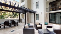 This inviting outdoor courtyard space is defined by its standout architectural centerpiece: a bold, dark-stained wood pergola that frames the patio and provides partial shade while maintaining an open, airy atmosphere. The slatted pergola structure casts striking linear shadows across the space, adding visual depth and a sense of sophistication to the overall design.

The patio flooring consists of large-format stone tiles laid in a classic pattern, offering a clean and durable surface that complements the contemporary aesthetic. Beneath the pergola, a generously arranged seating area features dark wicker lounge chairs with plush cream-colored cushions, creating a comfortable and stylish setting ideal for relaxation or social gatherings. A separate dining area with a dark table and chairs is positioned nearby, providing a dedicated space for outdoor meals.

Decorative potted bamboo and teal glazed planters add natural texture and a pop of color throughout the courtyard, softening the architectural lines. The space is nestled within a multi-story building complex with a crisp white stucco facade, and lush palm trees are visible in the background, suggesting a warm, tropical or subtropical setting. The courtyard's design balances modern elegance with functional outdoor living.
