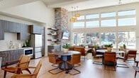 This single-floor community clubhouse delivers an impressive open-plan layout that seamlessly integrates a living room, dining area, and full kitchen into one cohesive, generously proportioned space. Soaring vaulted ceilings with coffered detailing amplify the sense of volume throughout, while an abundance of natural light pours in through floor-to-ceiling windows and clerestory windows that frame views of the outdoor pool and recreational area beyond.

The living area's centerpiece is a striking full-height stacked stone fireplace flanked by floating wood shelves, with a wall-mounted television above the firebox. A large sectional sofa, leather accent chairs, and woven ottomans create inviting conversation areas anchored by layered area rugs over light hardwood flooring. Rattan pendant lights add a coastal, organic warmth.

The dining zone features multiple round tables with leather cantilever chairs, ideal for communal gathering. The kitchen is equipped with stainless steel appliances, dark wood cabinetry with a textured wood-grain finish, and open shelving for display. A reception desk near the entry provides a functional welcome area with additional cabinetry. A vibrant floor-to-ceiling coral reef mural near the entrance adds a bold artistic focal point, completing the space's relaxed coastal-inspired aesthetic.