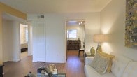 This single-story space is designed with an open, flowing layout that connects the main living area to the kitchen, creating a cohesive and functional floor plan. The living area features warm wood-style flooring that extends throughout, contributing to a welcoming and consistent aesthetic. Neutral-toned walls enhance the sense of brightness, complemented by soft ambient lighting from a table lamp that adds warmth to the space.

A louvered bifold closet door near the entryway provides practical storage while maintaining the clean, understated look of the interior. The hallway leads directly into the kitchen, which is visible in the background and features dark cabinetry, a sink positioned beneath a window that allows natural light to filter in, and track lighting overhead. The kitchen's warm wood flooring ties seamlessly into the rest of the space.

The living area is furnished with a light-colored sofa accented with patterned throw pillows, alongside decorative accessories that add character without overwhelming the space. Overall, the layout offers an efficient, comfortable environment with a neutral palette and practical design elements throughout.