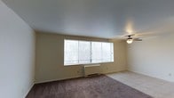 This single-floor space offers a clean and functional layout designed for comfortable everyday living. The main living area is a standout feature, characterized by a large, wide window that spans much of the exterior wall, allowing an abundance of natural light to fill the room and providing views of the surrounding greenery. Soft carpeted flooring runs throughout, adding warmth and comfort underfoot. A ceiling fan is mounted overhead, offering both practical air circulation and a subtle decorative touch to the smooth, neutral-toned ceiling. A wall-mounted heating unit sits beneath the window, ensuring year-round climate comfort. The walls are finished in a light, neutral palette that enhances the bright and airy atmosphere of the space. The overall ambiance is calm and understated, providing a blank canvas that lends itself well to a variety of interior styles and personal touches. The simplicity of the design prioritizes functionality, making it a practical and welcoming environment. Natural light remains a defining characteristic, giving the space an open, refreshing feel throughout the day.