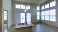 This single-floor space showcases a thoughtfully designed open-concept layout that emphasizes volume, light, and modern finishes. The soaring ceiling height is a defining architectural feature, creating an airy, expansive atmosphere that immediately draws the eye upward. A generous arrangement of large windows — including a striking row of clerestory windows positioned high on the walls — floods the interior with natural light throughout the day, offering sweeping outdoor views of the surrounding greenery.

At the heart of the space is a well-appointed kitchen island featuring elegant granite countertops with a deep single-basin sink, complemented by stylish pendant lighting that hangs gracefully from above. The kitchen flows seamlessly into the open living area, which is outfitted with durable wood-tone flooring and a ceiling fan to enhance comfort year-round.

A sliding glass door leads directly to an outdoor balcony, further blurring the boundary between interior and exterior living. The neutral color palette throughout — soft grays on the walls paired with light flooring — creates a clean, contemporary aesthetic. Corner windows in the living area amplify the sense of openness and provide multi-directional views. The overall ambiance is modern, airy, and inviting, making this a well-lit and functionally versatile space.