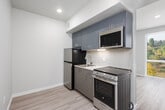 This single-floor unit features a thoughtfully designed layout that efficiently combines a kitchen, living area, and bathroom. The kitchen is outfitted with stainless steel appliances, including a refrigerator, electric range, and over-the-range microwave. Sleek gray cabinetry with brushed metal hardware provides ample storage, while a subway tile backsplash and light countertops contribute to a clean, contemporary aesthetic. Under-cabinet lighting enhances visibility and adds a polished finish to the space.

The main living area is bright and open, benefiting from a large floor-to-ceiling sliding glass door that invites abundant natural light and frames views of the surrounding urban and green landscape. A wall-mounted split HVAC unit ensures year-round climate control. Light gray hardwood-style flooring runs throughout, lending a cohesive, modern feel to the entire unit.

The bathroom is efficiently appointed with a single-sink vanity featuring a white countertop and storage cabinetry below, a full-width mirror, and a combined shower and toilet area. Neutral wall tones and chrome fixtures give the bathroom a clean, functional character. Overall, the unit presents a streamlined, contemporary living environment suited for efficient, comfortable everyday use.