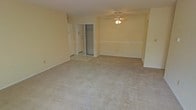 This single-floor residential space offers a clean and functional layout designed for comfortable everyday living. The main living area is generously sized, featuring wall-to-wall neutral-toned carpet that adds warmth and softness underfoot. A ceiling fan with an integrated light fixture is centrally positioned overhead, providing both air circulation and ambient lighting throughout the room. The walls are finished in a light, neutral tone that enhances the sense of openness and brightness within the space. A chair rail-style dividing line runs along the walls, adding a subtle architectural detail. Adjacent to the main living area is a tiled entryway, which transitions smoothly from the carpeted interior, providing a practical and easy-to-clean surface near the entrance. A closet with sliding doors is conveniently located near the entry, offering accessible storage space. The overall ambiance of the space is bright, open, and move-in ready, with a simple and versatile interior that allows for a variety of furniture arrangements and personal styling choices. The layout is thoughtfully designed to maximize usable floor space while maintaining an uncluttered, airy feel throughout.