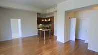 This single-floor space is defined by an open-concept layout that connects the living area to the kitchen, creating a cohesive and functional environment. Warm-toned hardwood flooring extends throughout, lending a consistent and inviting character to the space.

The kitchen is positioned toward the rear and features rich dark wood cabinetry that reaches toward the ceiling, providing ample storage. A decorative mosaic tile backsplash adds visual interest along the walls, complementing the stainless steel appliances, including a range and over-the-range microwave. A built-in kitchen island with a light-toned countertop and chrome legs serves as a natural dividing point between the kitchen and the open living area, functioning equally well as a prep surface or casual dining spot.

Above the island, a set of three pendant lights hangs from a track-mounted rail system, casting warm, focused light over the workspace. The open living area is generous in scale, with clean white walls and simple architectural transitions between spaces. A partial wall with a wide opening frames the kitchen area, adding subtle definition without closing off the layout.