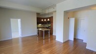 This single-floor space is defined by an open-concept layout that connects the living area to the kitchen, creating a cohesive and functional environment. Warm-toned hardwood flooring extends throughout, lending a consistent and inviting character to the space.

The kitchen is positioned toward the rear and features rich dark wood cabinetry that reaches toward the ceiling, providing ample storage. A decorative mosaic tile backsplash adds visual interest along the walls, complementing the stainless steel appliances, including a range and over-the-range microwave. A built-in kitchen island with a light-toned countertop and chrome legs serves as a natural dividing point between the kitchen and the open living area, functioning equally well as a prep surface or casual dining spot.

Above the island, a set of three pendant lights hangs from a track-mounted rail system, casting warm, focused light over the workspace. The open living area is generous in scale, with clean white walls and simple architectural transitions between spaces. A partial wall with a wide opening frames the kitchen area, adding subtle definition without closing off the layout.