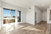 This single-floor unit showcases a clean, contemporary design characterized by crisp white walls, light wood-tone flooring, and recessed lighting throughout. A bright hallway connects the main living areas, creating a logical and efficient flow from entry to living space. The kitchen is a functional highlight, featuring warm wood-grain cabinetry extending to the ceiling, white quartz-style countertops, and a full suite of stainless steel appliances including a refrigerator, electric range, built-in microwave, and dishwasher — all set against a white tile backsplash. The open layout allows the kitchen to integrate naturally with the adjacent living area, which benefits from natural light streaming in from large windows. The bedroom is bright and airy, with a generously sized window framing views of the surrounding treetops and neighborhood below. The bathroom is finished in an all-white palette, featuring a soaking tub with an overhead shower, subway-style tile surround, brushed nickel fixtures, and a small window for natural ventilation. Throughout the unit, the consistent use of light tones and modern finishes creates a cohesive, well-lit, and welcoming atmosphere.