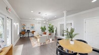 Located at 3661 Airport Blvd in Mobile, Alabama, this single-floor commercial space offers a thoughtfully designed, open-plan layout that balances functionality with modern aesthetic appeal. The interior is finished with warm hardwood-style flooring throughout, crisp white walls, and elegant crown molding that adds a refined architectural touch. A decorative white column anchors the central area, providing visual structure without dividing the open flow of the space.

The lounge area serves as the welcoming focal point, featuring a curved cream sofa, accent chairs, and a bohemian-style area rug, all illuminated by striking sputnik-style chandeliers that lend a contemporary flair. Lush fiddle-leaf fig trees and tropical palms are positioned throughout, adding vibrant, natural character.

Adjacent to the lounge, a dedicated workspace with a desk and office chairs provides a functional area for productivity. A round dining table with seating occupies another zone near the entry, complemented by framed wall art and a console table.

The kitchen is well-appointed with dark espresso cabinetry, a mosaic tile backsplash, granite countertops, and stainless steel appliances including a refrigerator, range, and dishwasher. A large island with bar seating adds both prep space and casual dining versatility. Multiple windows throughout ensure ample natural light, creating a bright and inviting atmosphere.