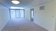 This single-floor space offers an open and airy layout defined by its generous floor-to-ceiling windows along one wall, allowing abundant natural light to flood the interior. The expansive main living area is finished with neutral wall-to-wall carpeting, contributing to a calm, understated atmosphere. A angled ceiling detail near the window wall adds a subtle architectural interest, while a flush ceiling light fixture provides overhead illumination. The crisp white walls throughout maintain a clean, bright aesthetic. A wall-mounted HVAC vent ensures climate control convenience. An open doorway connects the main living area to an adjacent room, which also benefits from natural light via its own window, suggesting a versatile secondary space. The overall design is straightforward and functional, with minimal ornamentation, making it well-suited for a variety of uses. Neutral finishes and an uncluttered layout give the space a flexible, move-in-ready quality that can easily accommodate diverse furnishing arrangements.