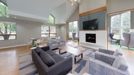 This well-appointed single-floor community space showcases a thoughtful modern design with vaulted ceilings that create an open, airy atmosphere throughout. The main lounge area serves as the centerpiece, featuring a striking fireplace framed by wood accents and a patterned tile surround, a wall-mounted television, and comfortable gray upholstered seating arranged on a geometric area rug. Soaring angled ceilings with clerestory windows flood the space with natural light, offering views of surrounding greenery.

Adjacent to the lounge, a casual seating nook provides an inviting transitional area with a mix of high-top bar seating and green accent chairs, creating an informal gathering spot. The dining area is bright and welcoming, furnished with wood-topped tables paired with modern white chairs, all set against warm hardwood flooring and surrounded by large wood-trimmed windows overlooking mature trees.

The kitchen is sleek and functional, anchored by an expansive granite-look countertop bar, warm wood cabinetry, stainless steel appliances, and a subway tile backsplash. Pendant lighting with yellow-accented interiors adds a stylish touch. Throughout the space, consistent hardwood flooring, soft gray walls, and an abundance of natural light contribute to a cohesive, welcoming environment well-suited for communal gatherings.