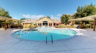 This expansive outdoor amenity area serves as a central gathering hub, highlighted by a generously sized swimming pool with crystal-clear blue water and smooth stone coping. Adjacent to the main pool sits a separate spa, providing a dedicated space for relaxation. The surrounding sun deck is finished in light-toned pavers and is well-appointed with numerous sling-style lounge chairs, side tables, and colorful cantilever umbrellas in red and green, offering ample shaded and open-air seating options.

A pergola structure with slatted roofing extends along one side of the pool area, supported by warm-toned columns and framed by raised brick planters overflowing with ornamental grasses and seasonal flowers. This shaded zone features additional table-and-chair seating, making it ideal for outdoor dining or socializing. A secondary patio near the community clubhouse building provides further seating beneath mature shade trees, complemented by colorful potted plants and manicured landscaping. The brick-accented clubhouse building with arched windows anchors the space architecturally, while well-maintained greenery throughout creates a resort-like ambiance under abundant natural sunlight.
