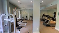 This fitness center offers a well-organized and welcoming environment for exercise and wellness. The space is finished with soft sage-green walls and neutral carpeted flooring, creating a calm, focused atmosphere conducive to working out. Recessed lighting throughout the ceiling ensures consistent, bright illumination across the entire floor, while a ceiling fan provides additional air circulation for comfort during workouts. The layout is thoughtfully divided by a structural column, creating distinct zones for strength training and cardiovascular exercise. The strength training area is equipped with multiple white-framed cable and resistance machines, including seated cable stations and adjustable weight machines. Natural light filters in through a window along one wall, adding to the open and airy feel of the space. The cardio zone features upright exercise bikes and an elliptical trainer, offering a range of low-impact aerobic options. White baseboards and clean wall finishes give the room a polished, well-maintained appearance. An emergency exit sign is visible, indicating compliance with safety standards. Overall, this fitness center combines functionality with a tidy, comfortable design suitable for a broad range of exercise routines.