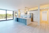 This contemporary single-floor residence showcases a thoughtfully designed open-concept layout that seamlessly connects the living and kitchen areas. The kitchen is a standout feature, centered around a spacious island with striking blue-gray cabinetry and a pristine white quartz countertop. White upper cabinetry with gold hardware, a professional stainless steel range with hood, a full-size refrigerator, and a dedicated sink area complete the culinary space. Two geometric brass pendant lights anchor the kitchen, adding a warm, upscale ambiance. Light-toned large-format tile flooring runs throughout the entire residence, creating a clean and cohesive aesthetic. Multiple floor-to-ceiling sliding glass doors open to generous outdoor balconies, flooding the interior with abundant natural light and offering lush treetop views. The bathrooms are finished with white vanities accented by gold hardware; one features a walk-in marble-tiled shower with a sleek glass sliding door. Ample storage is provided through multiple well-organized walk-in closets outfitted with custom white shelving systems and gold hanging rods. A powder room with matching finishes offers additional convenience. The overall design is bright, modern, and cohesive, with gold accents tying every space together.